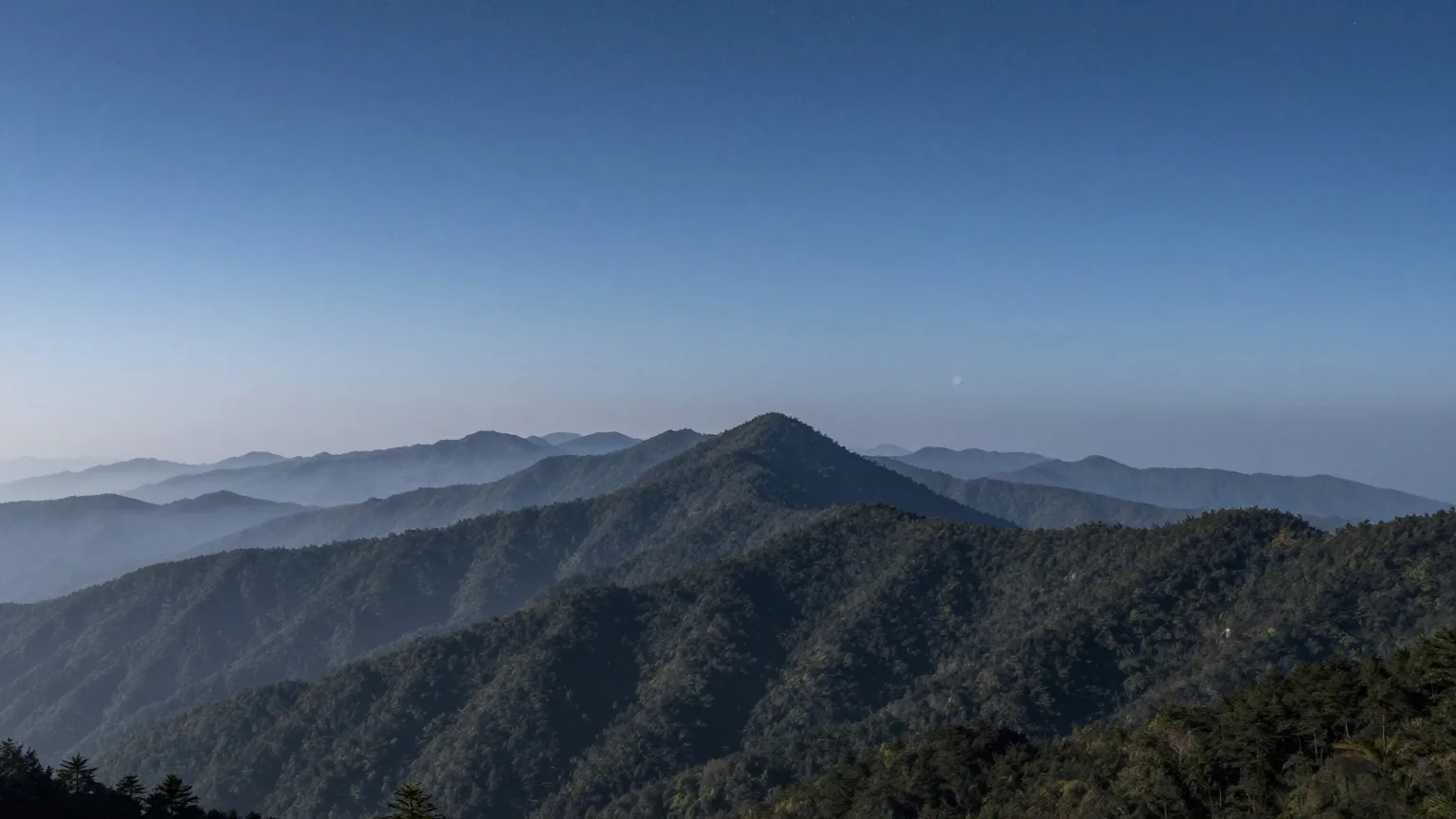 Bewaldete Bergrücken unter klarem Sternenhimmel, Nebel in den Tälern und eine schmale Mondsichel über dem Horizont