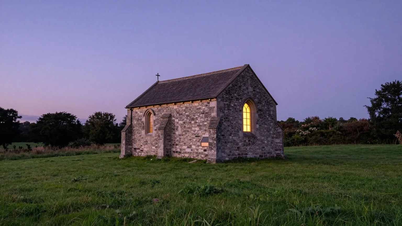 Kleine Steinkapelle in einer gemähten Wiese in der Abenddämmerung, ein Fenster leuchtet warm, am violetten Himmel erscheinen die ersten Sterne