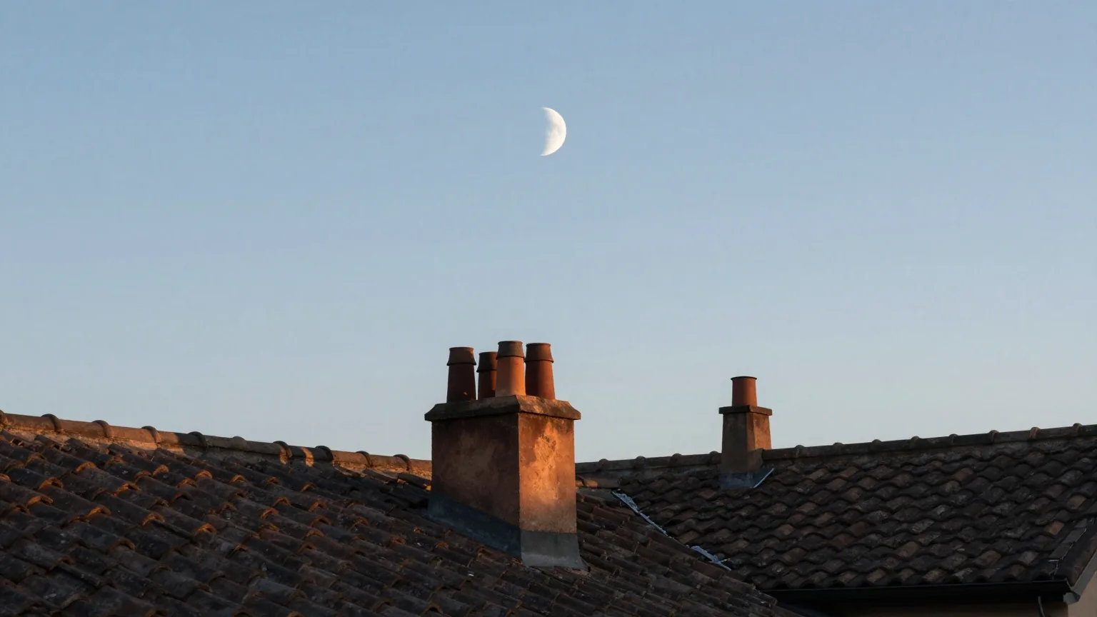 Blasse Mondsichel am hellblauen Morgenhimmel über stillen Ziegeldächern, die ersten Sonnenstrahlen treffen die Schornsteine