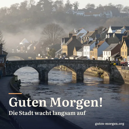 Historische Steinbrücke über einem breiten Fluss, Altstadthäuser im ersten Sonnenlicht, leichter Nebel über dem Wasser; Schrift: "Guten Morgen! Die Stadt wacht langsam auf"
