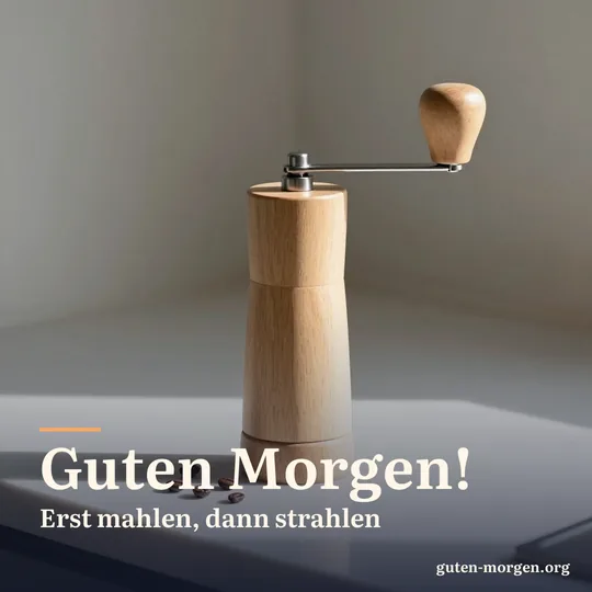 Alte hölzerne Handkaffeemühle mit ein paar Bohnen auf der Küchenarbeitsplatte, ein Sonnenstrahl fällt darauf; Schrift: „Guten Morgen! Erst mahlen, dann strahlen“