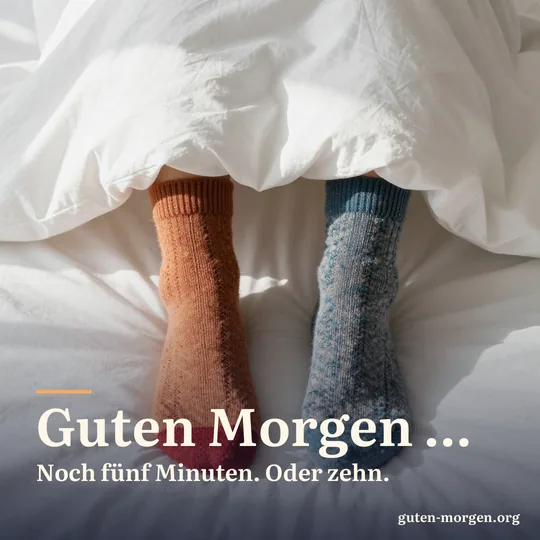 Zwei Füße in unterschiedlichen bunten Wollsocken schauen unter einer dicken weißen Bettdecke hervor; Schrift: "Guten Morgen … Noch fünf Minuten. Oder zehn."